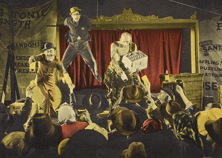 A stage, with a man dangling from a suspended bar. Two other men on the stage give bottles to the audience.