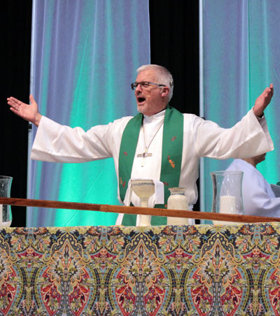 A white man with gray hair and glasses, wearing a long white robe and a green stole, with his arms outstretched.